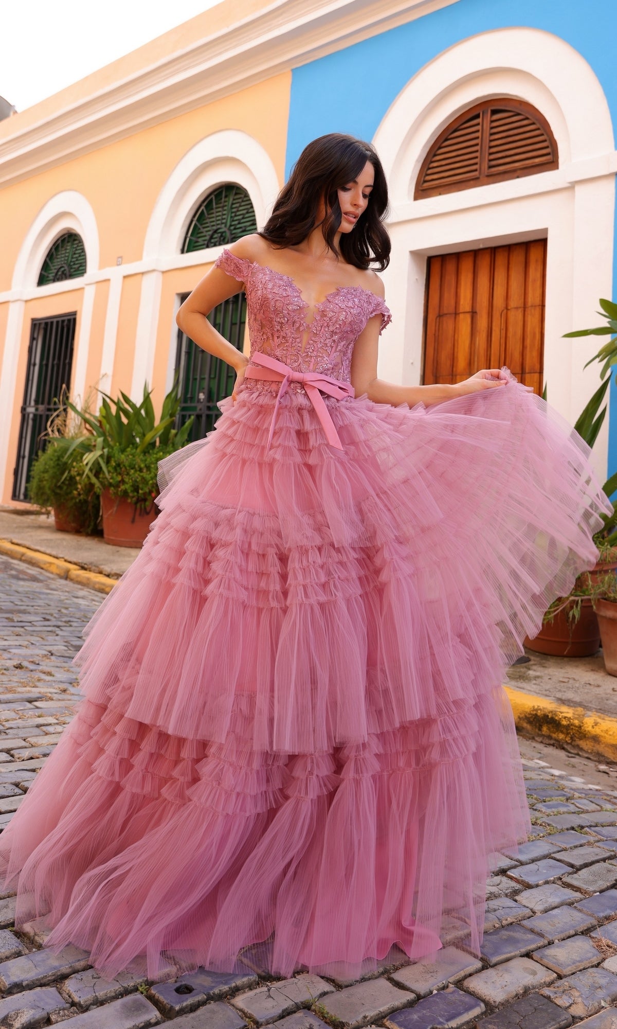 A woman in the Nox Anabel Designer Ruffled Prom Ball Gown E1293, with layered pink tulle and an off-the-shoulder design, stands on a cobblestone street framed by colorful buildings and lush green plants.